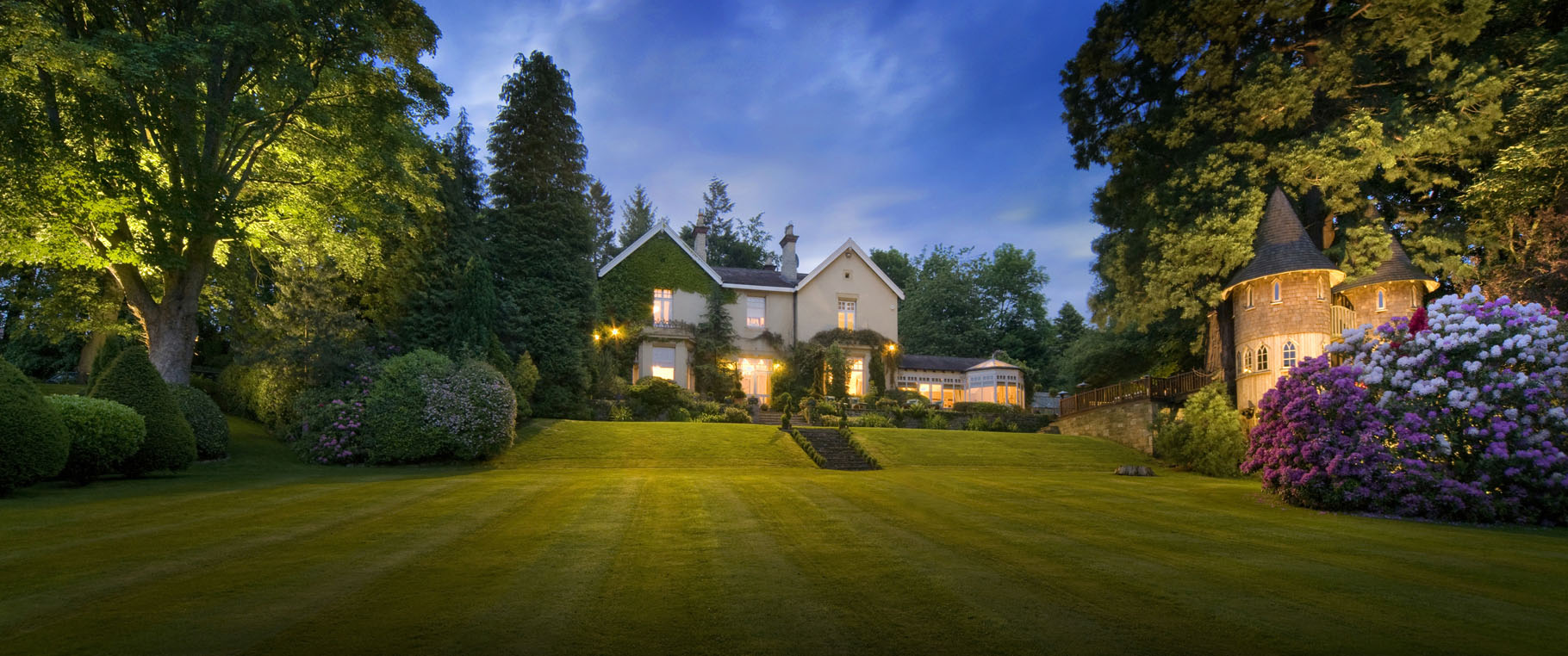 Architectural exterior of property set into gardens with treehouse which are illuminated at dusk