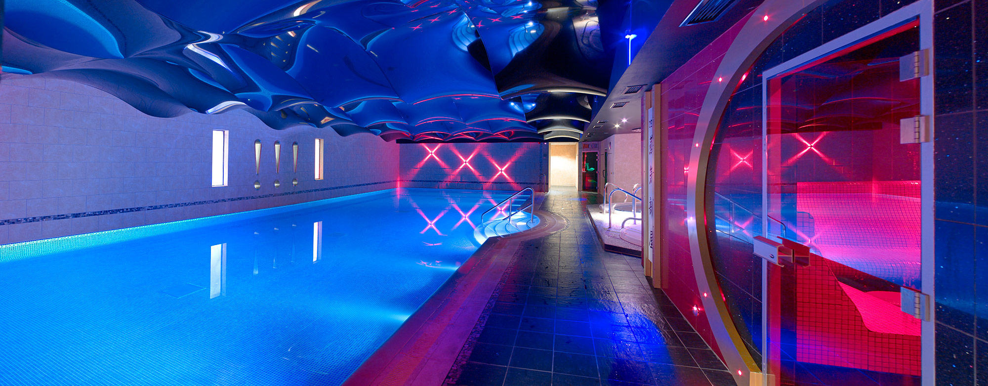 Illuminated modern indoor swimming pool in with mirrored ceiling and blue and pink feature lighting