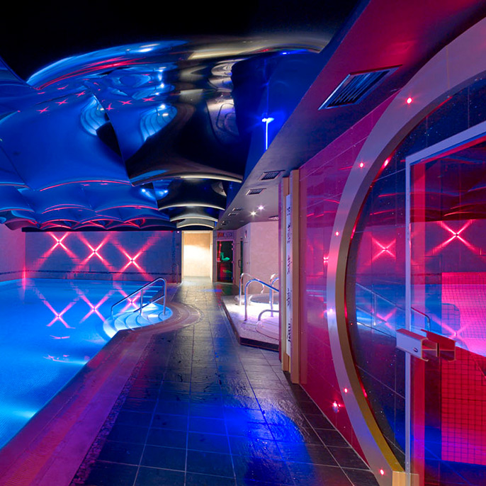 Illuminated modern indoor swimming pool in with mirrored ceiling and blue and pink feature lighting