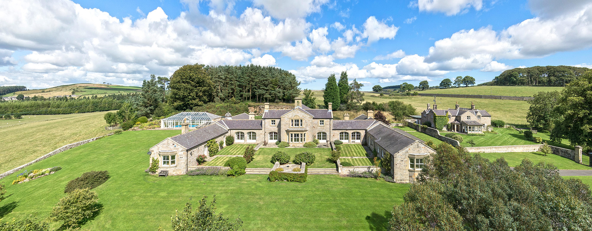 A view over a countryside property with bue sky fluffy clouds, trees and landscape